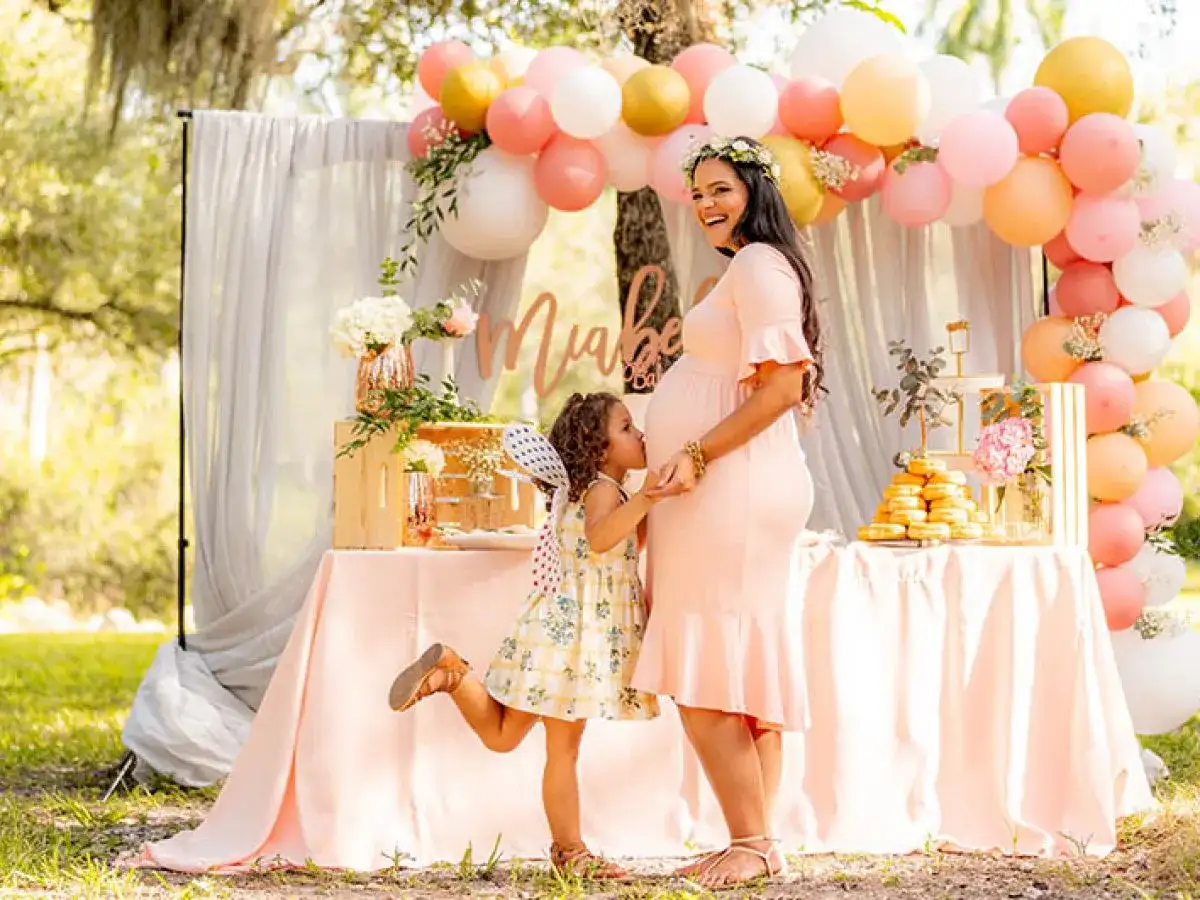 Uśmiechnięta ciężarna kobieta i mała dziewczynka przy stole z balonami i słodkościami. Wspaniałe atrakcje na baby shower w plenerze.