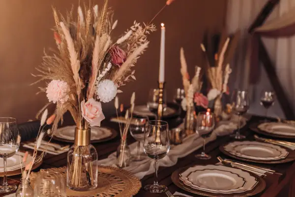 Eleganckie inspiracje weselne: boho bukiet z traw, róż i pampasów na drewnianym stole z kryształowymi kieliszkami i złotymi naczyniami.