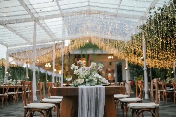Elegancki wystrój sali weselnej pod baldachimem z tysięcy światełek. Stół z bukietem kwiatów i drewniane krzesła tworzą magiczną atmosferę.